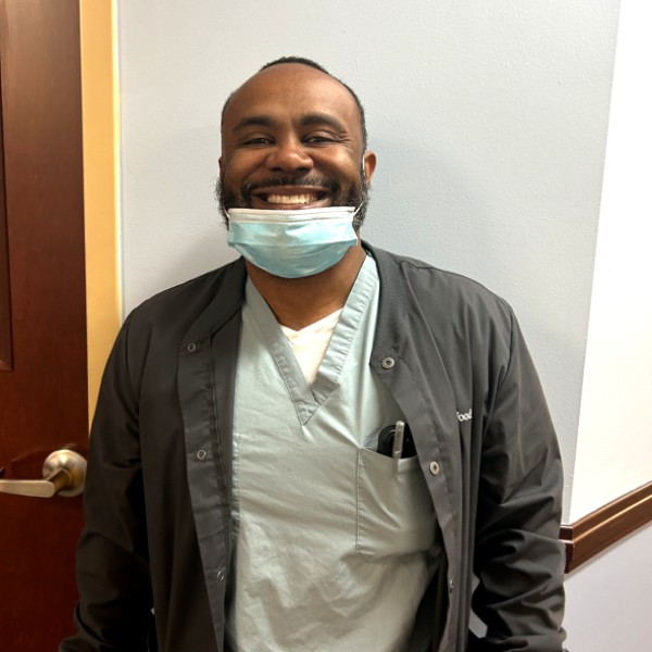 Smiling man in light green medical scrubs under a dark jacket mask pulled under his chin standing by a doorway in a clinic setting Smiling man in light green medical scrubs under a dark jacket, mask pulled under his chin, standing by a doorway in a clinic setting.